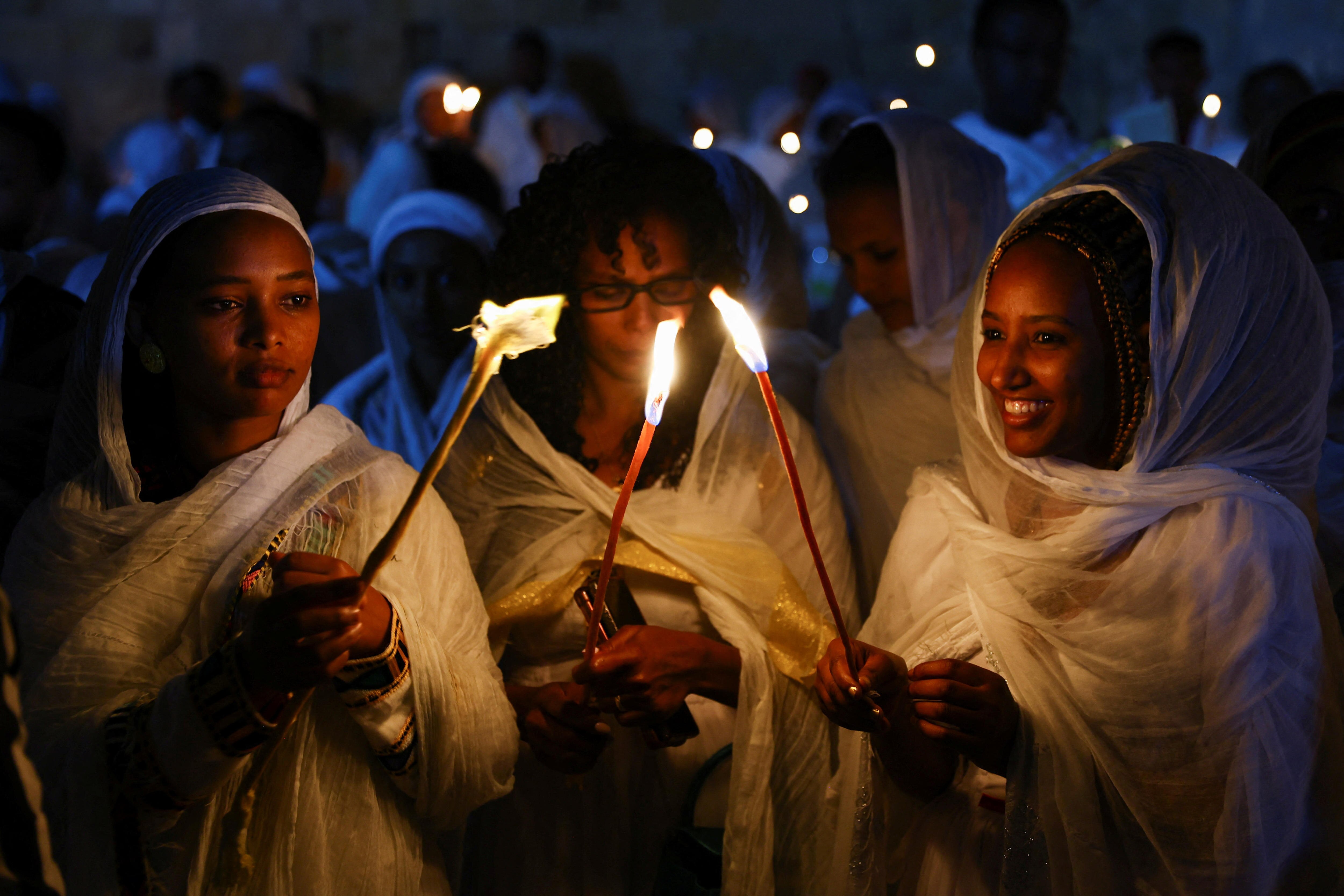 Three women stand together holding candles which glow with bright orange flames.