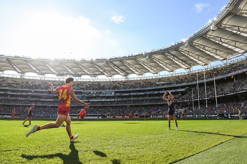 An AFL footballer runs in to kick for goal from near the boundary in the right forward pocket as a defender stands the mark.