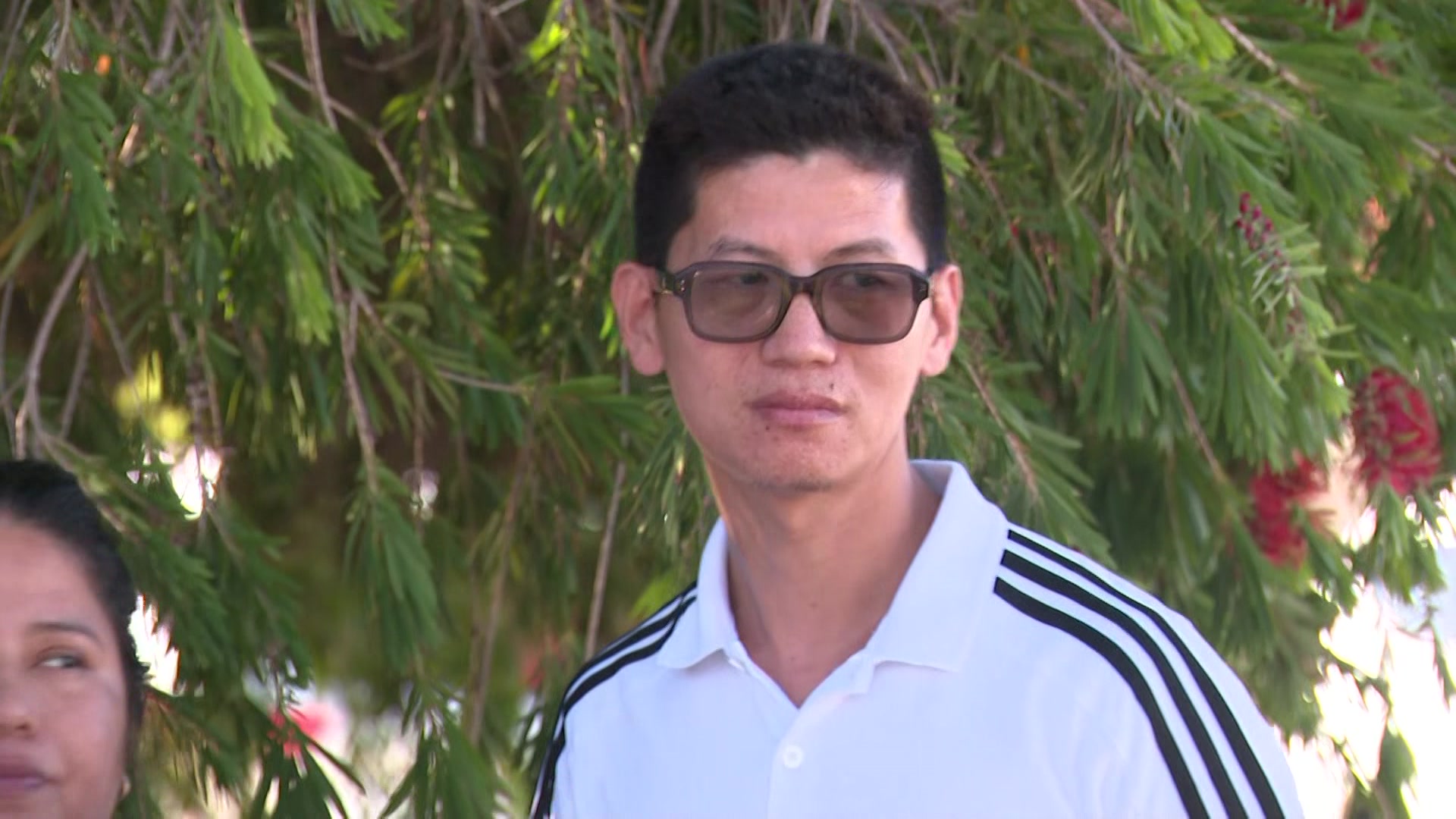 Un hombre con gafas se para frente a un árbol.