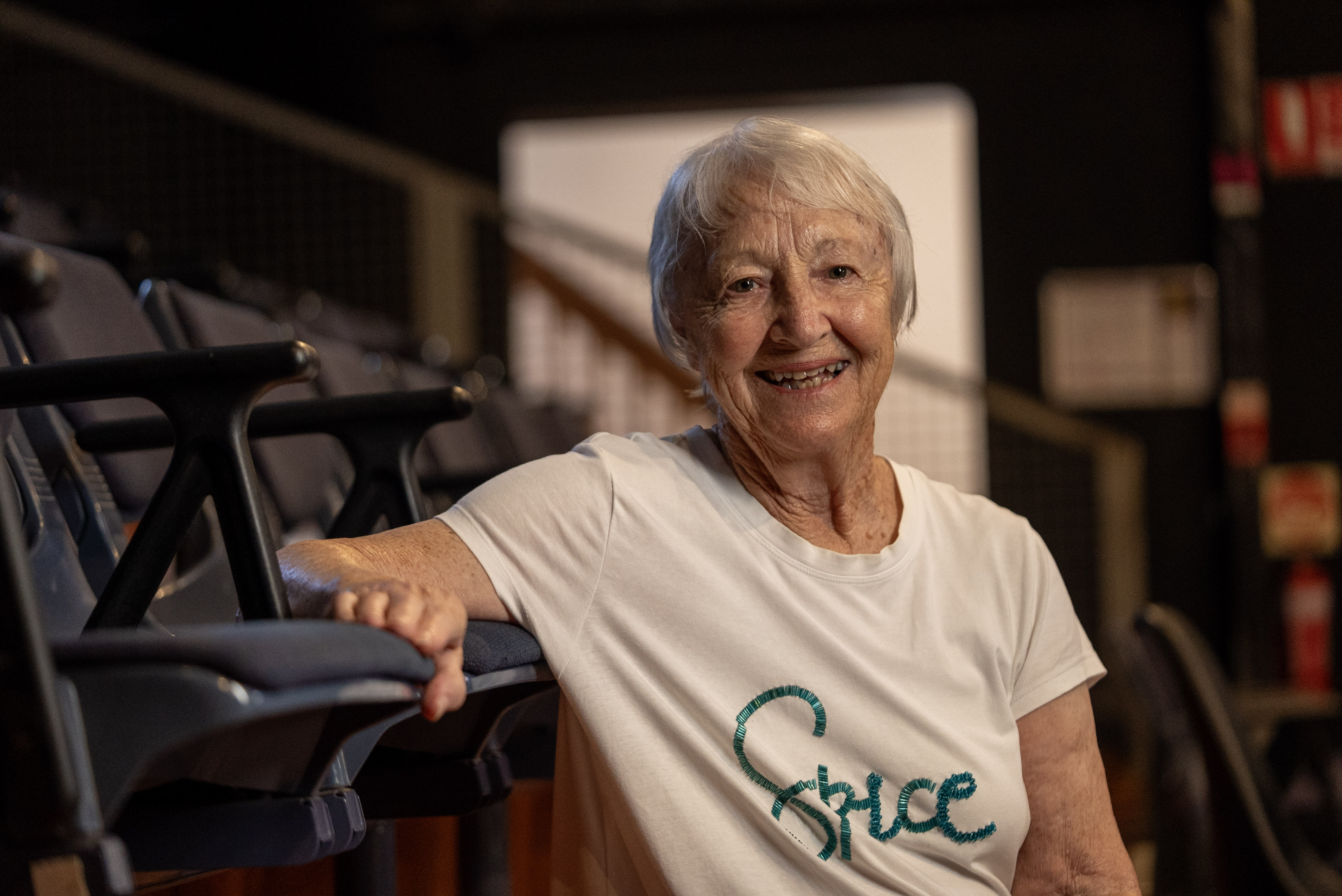 An older woman in a while shirt with short grey hair sits back in a chair in a theatre.