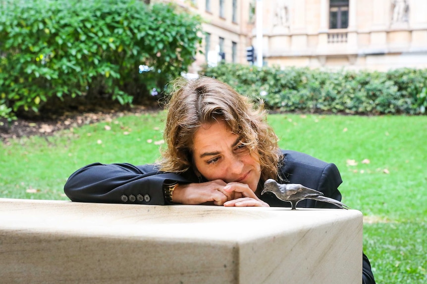 Mid-shot of artist Tracey Emin resting on a sandstone birdbath in a park, closely inspecting a bronze bird-like sculpture.