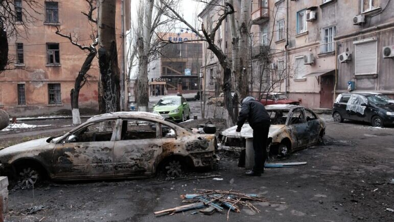 A man stands next to two burnt out cars 
