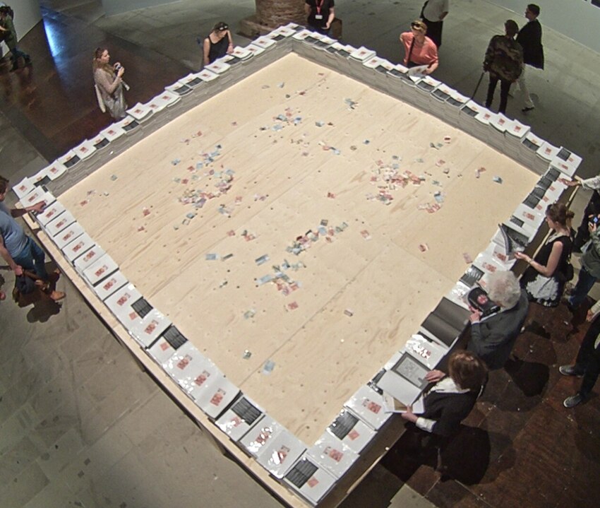 People stand around a square table, perusing books that are stacked around its edges. Euros are scattered in the centre