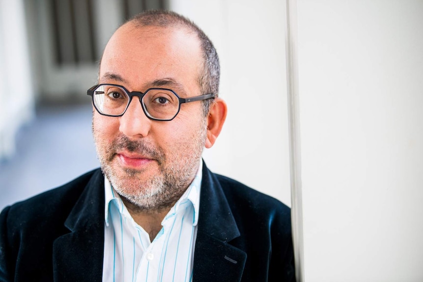 Close-up of man with balding head, wearing glasses and dark teal velvet jacket with blue-on-white striped shirt.