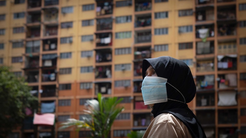 A woman in a hijab wears a mask in front of a high-rise building.