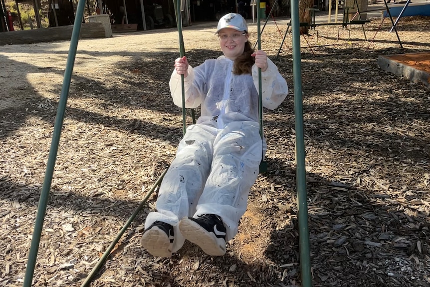 A young woman in a cap, glasses and with a painting suit over her clothes, smiles as she sits on a swing.