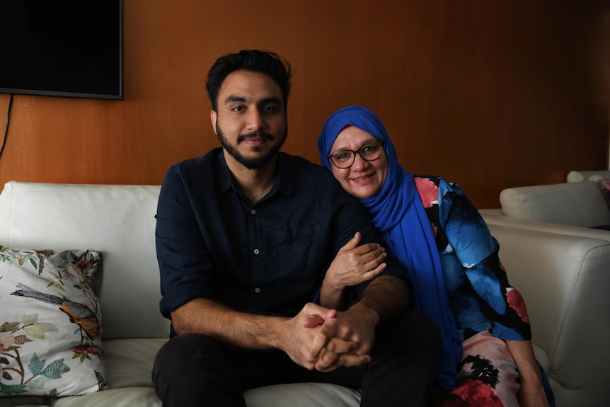 Woman wearing glasses, an electric blue headscark and bright dress sits on a couch next to a man who wears black shirt, pants