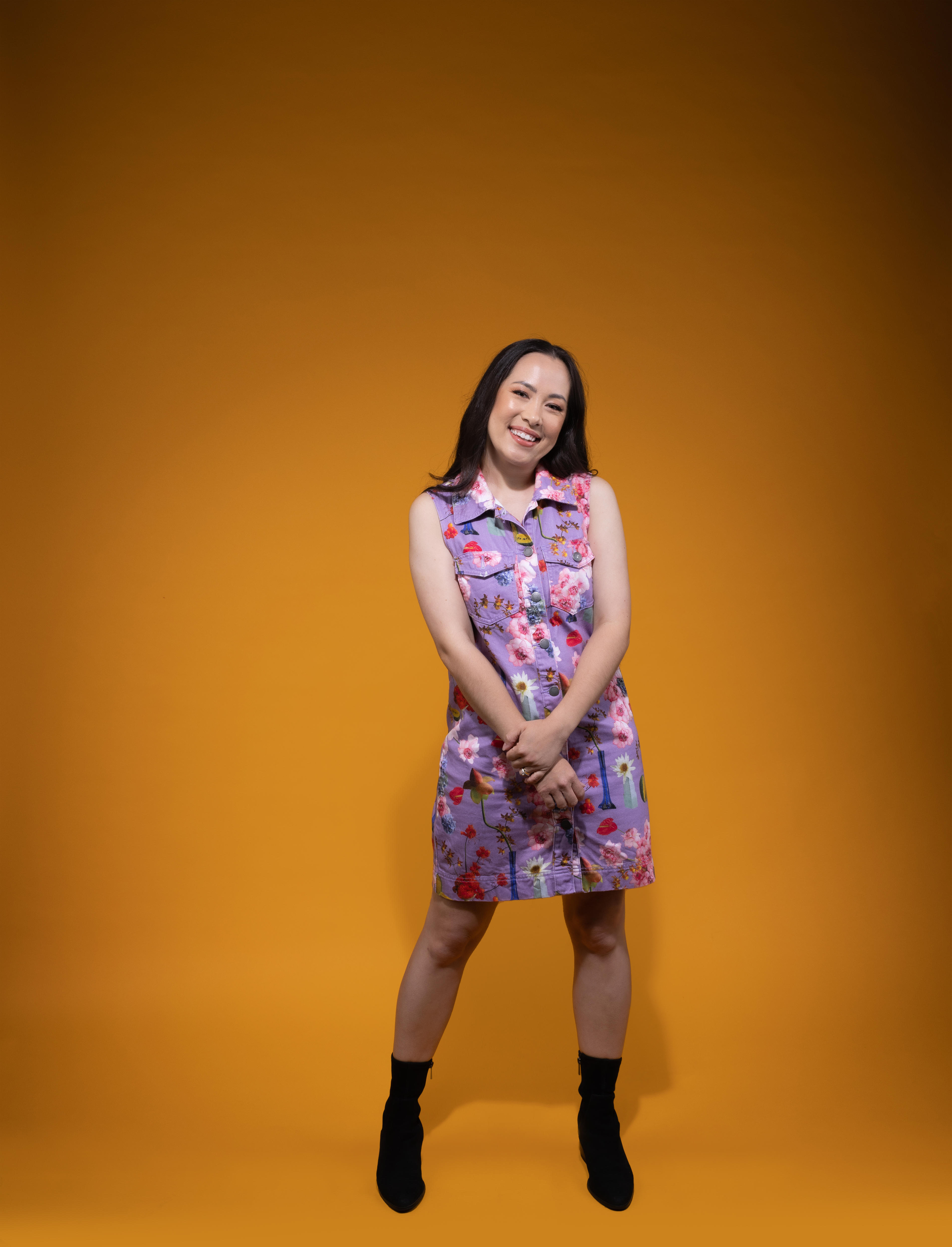 Asian Australian woman with long dark hair wears purple dress with colourful splodges while standing against a yellow backdrop.