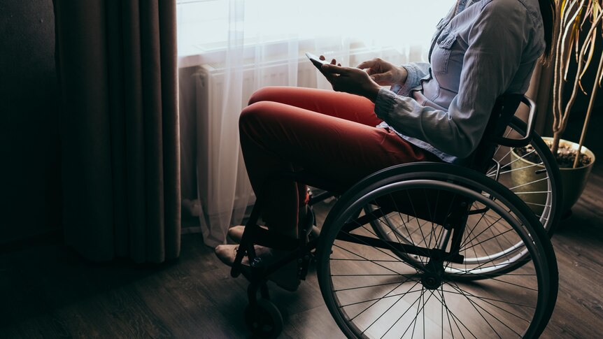 A young woman sitting in a wheelchair by a window is using a smartphone, the lighting in the room is shadowy and dark. 