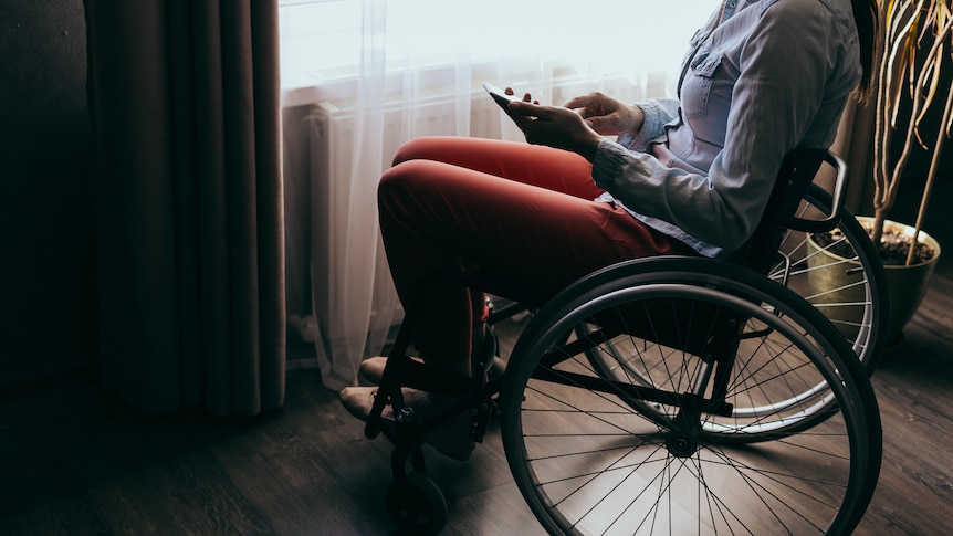 A woman in a wheelchair sitting by a window using a smartphone.