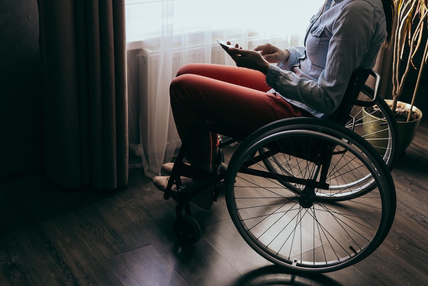A young woman sitting in a wheelchair by a window is using a smartphone, the lighting in the room is shadowy and dark. 