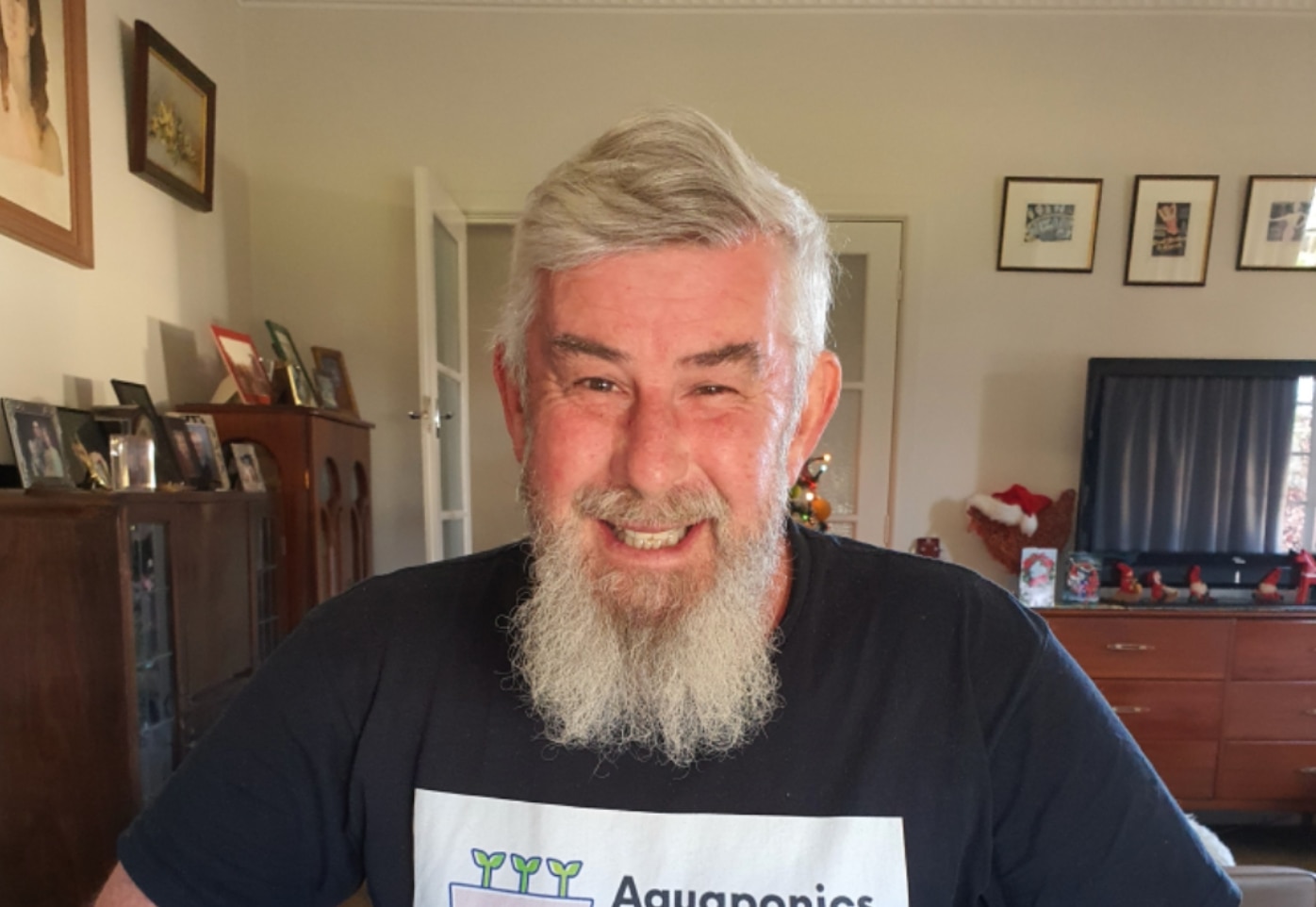 A man with white hair and a white beard smiles. He wears a black T-shirt.