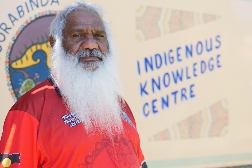 A man with grey hair and bushy grey beard stands in front a wall that says Indigenous Knowledge Centre.