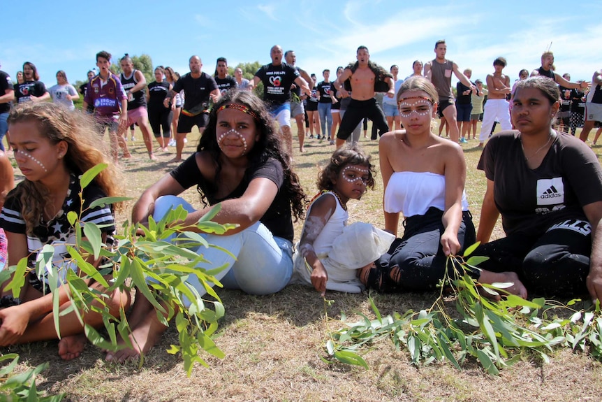 Young corroboree participants seated outside with haka dancers in the background.