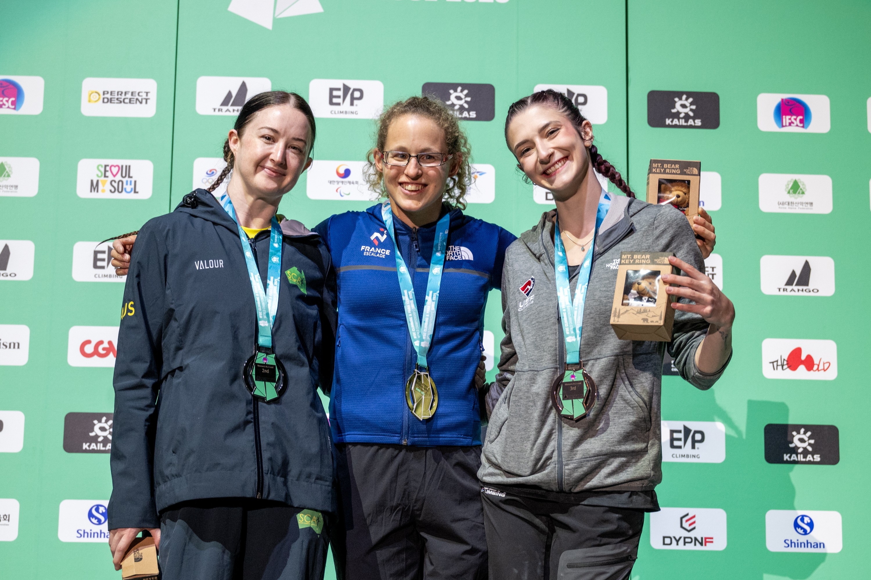 As alpinistas Sarah Larcombe, Lucie Jarrige e Daliya Hansen estão de braços dados no pódio enquanto usam suas medalhas.