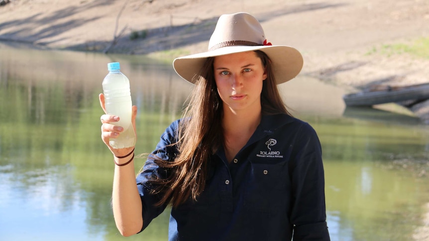 A woman with long brown hair and wearing a wide brim hat holds up a bottle of murky water