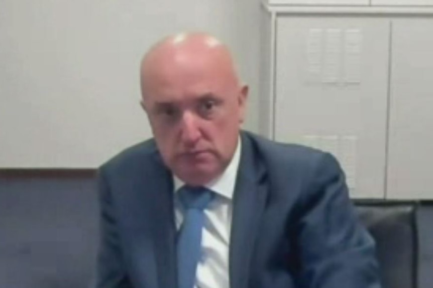 A bald man in a suit sitting in front of a microphone in an official looking setting.