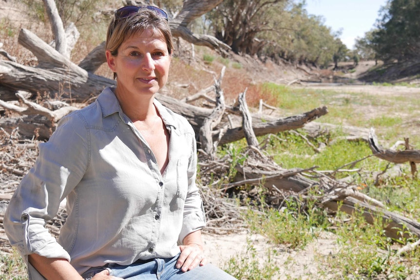 A woman sitting down in a dry riverbed littered with the skeletal remains of trees.