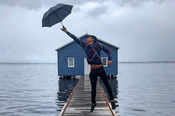 A man jumps while holding an umbrella on a jetty leading up to a blue boathouse.