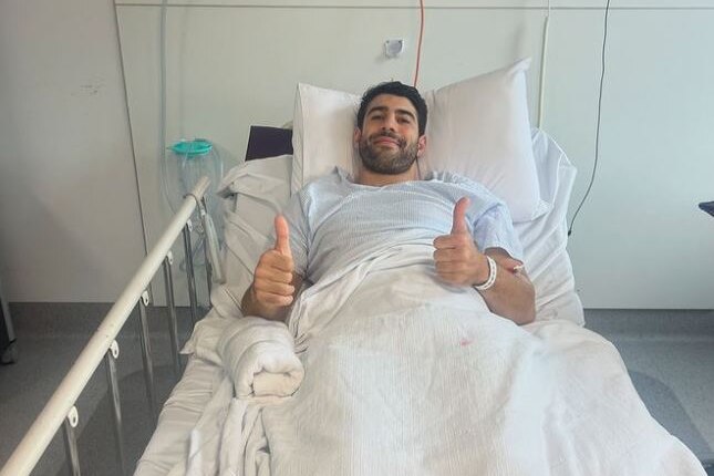 An AFL player lies in a hospital bed giving two thumbs up to the camera.