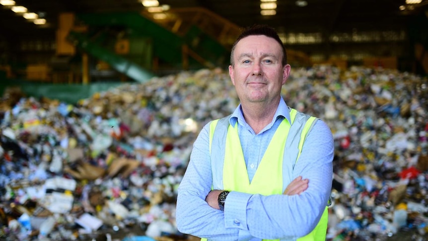 Man in a blue shirt and high vis vest smiling at the camera. Background is piles of rubbish.