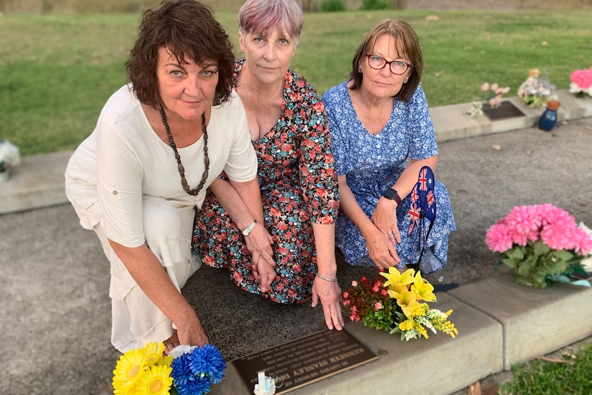 Three sisters kneeling in front of their father's gravestone, with flowers placed on the ground.