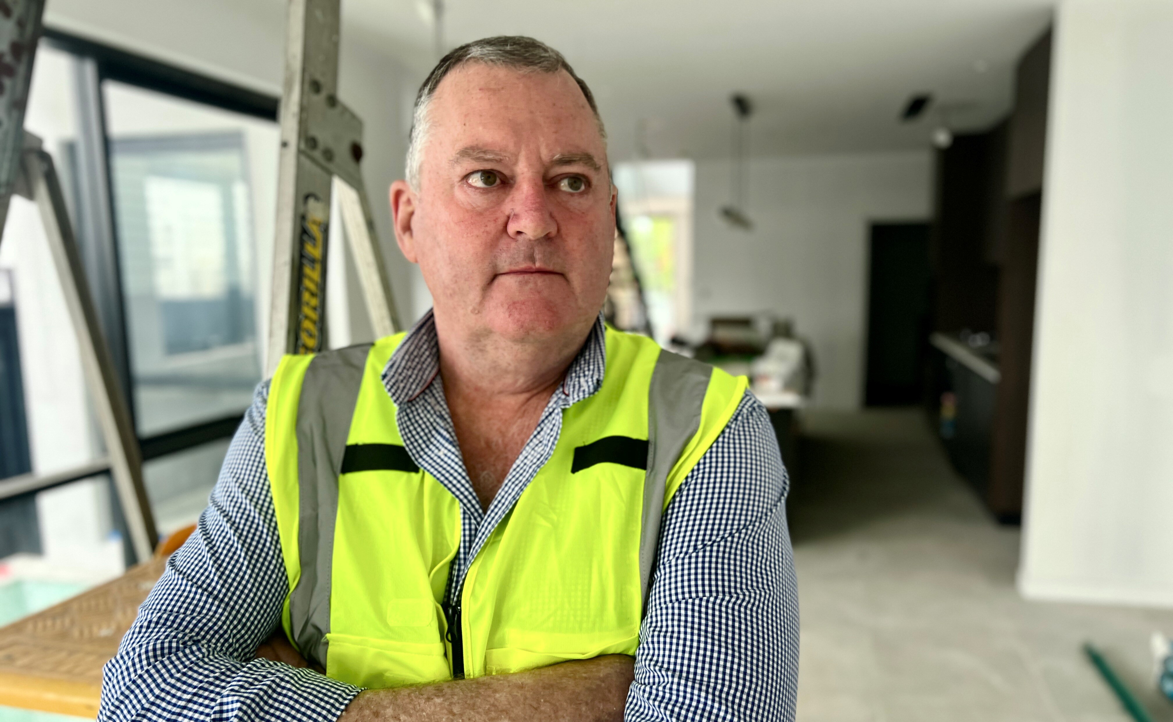 Middle-aged Scott is serious, looks off camera, wears high-viz jacket over check shirt, stands in room under construction.