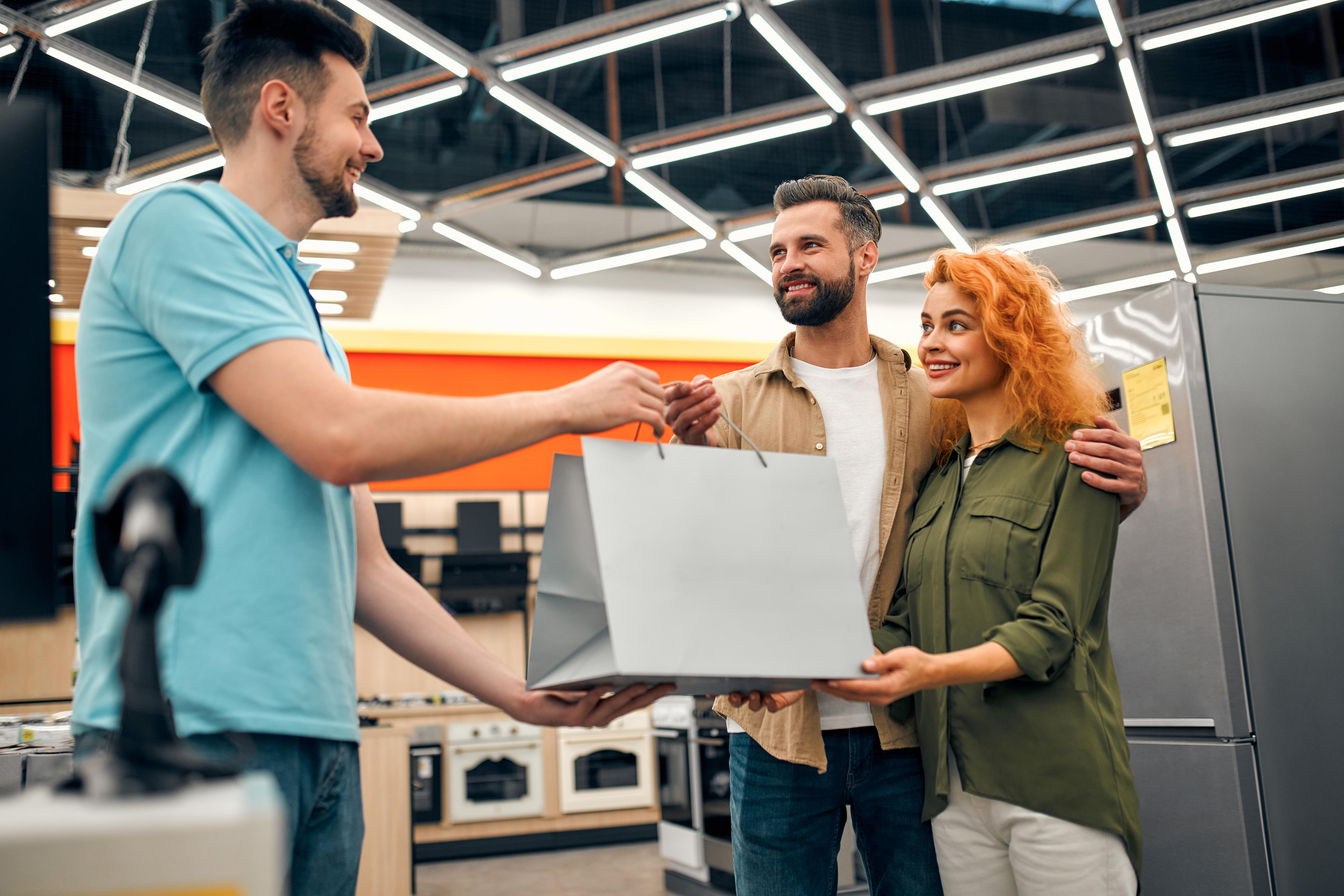 A couple stand smiling in an electronics retailer as a salesman hands them a bag