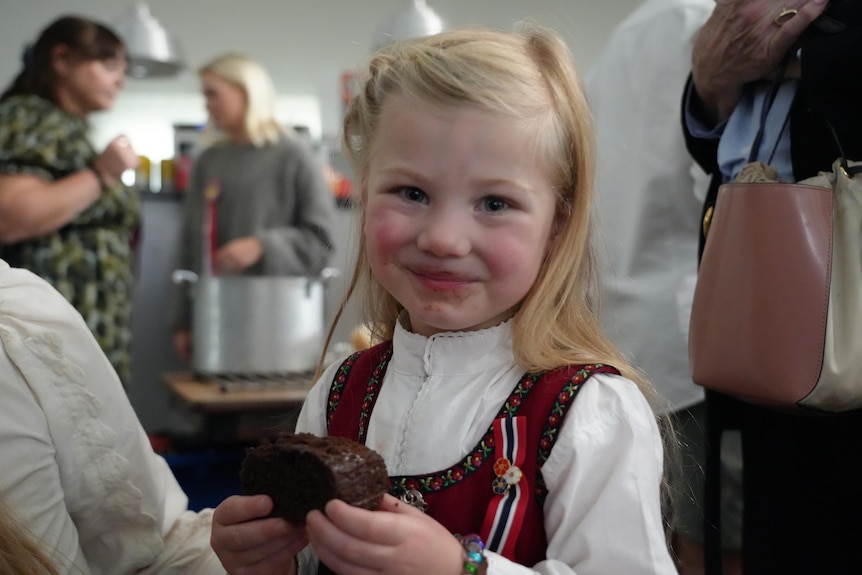 A little girl with blonde hair and dressed in traditional Norwegian clothing eats a piece of chocolate cake.
