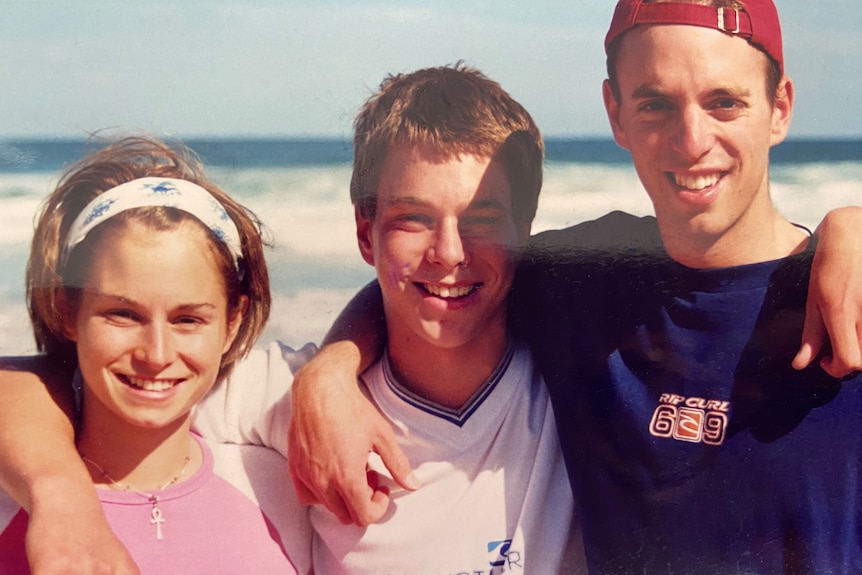 Two teenage boys and a teenage girl with arms around each other's shoulders at the beach
