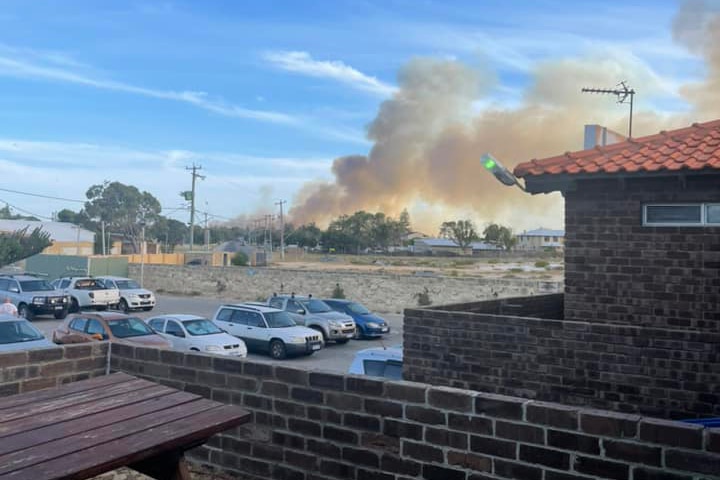 A bushfire burns against a blue sky with cars parked in the foreground