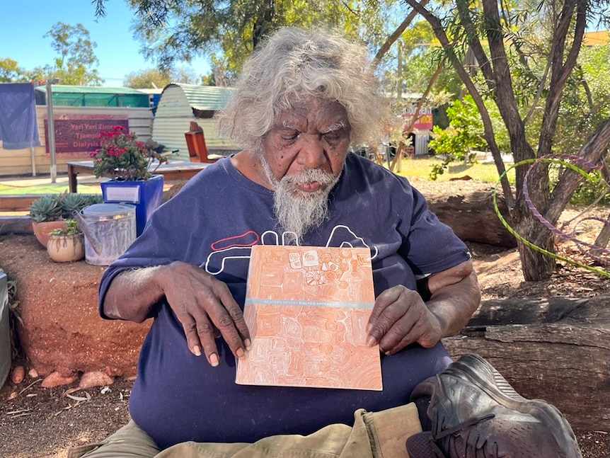 An Indigenous man sits holding a brochure with art on it. He has grey hair and looks down at the picture. He sits under a tree.