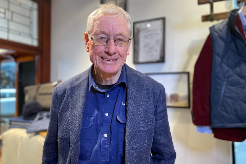 An older man with glasses stands in front of a shop wall, with a clothes rack to his right