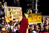 A crowd of people protesting, with some people holding signs saying 'not the Queen's land' and holding flags.