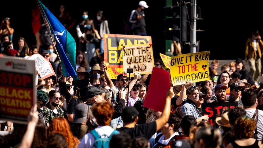 A crowd of people protesting, with some people holding signs saying 'not the Queen's land' and holding flags.