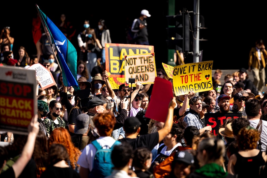 A crowd of people protesting, with some people holding signs saying 'not the Queen's land' and holding flags.