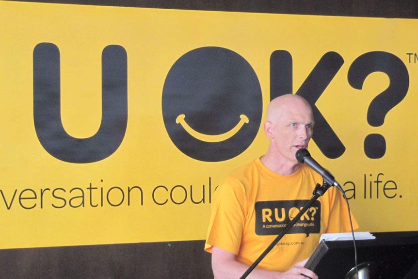 A bald Gavin stands at a podium in front of an R U OK? banner