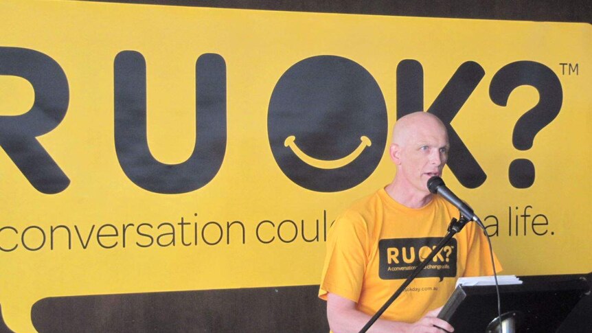 A bald Gavin stands at a podium in front of an R U OK? banner