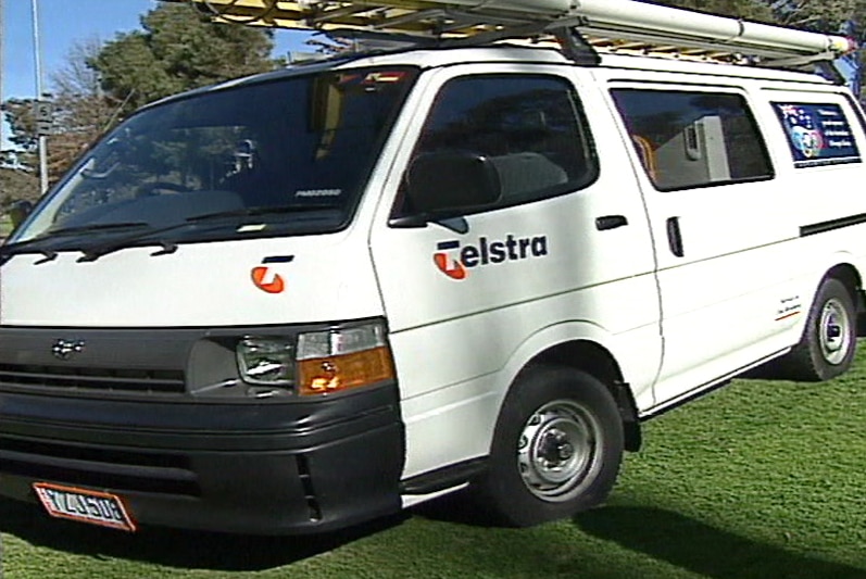 A Telstra work van from the mid-1990s with a ladder on the roof parked on a lawn.