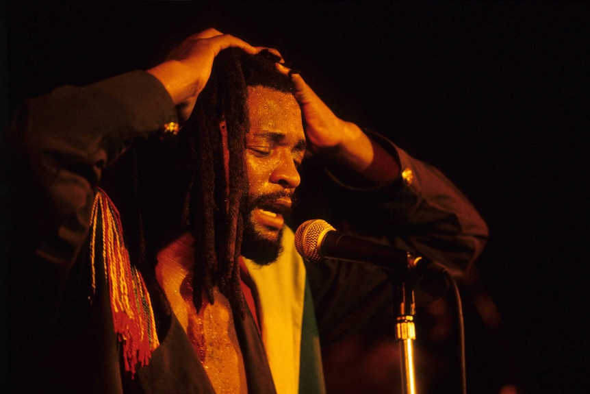 Midshot of a performer on stage, with his arms on top of his head, singing into the microphone.