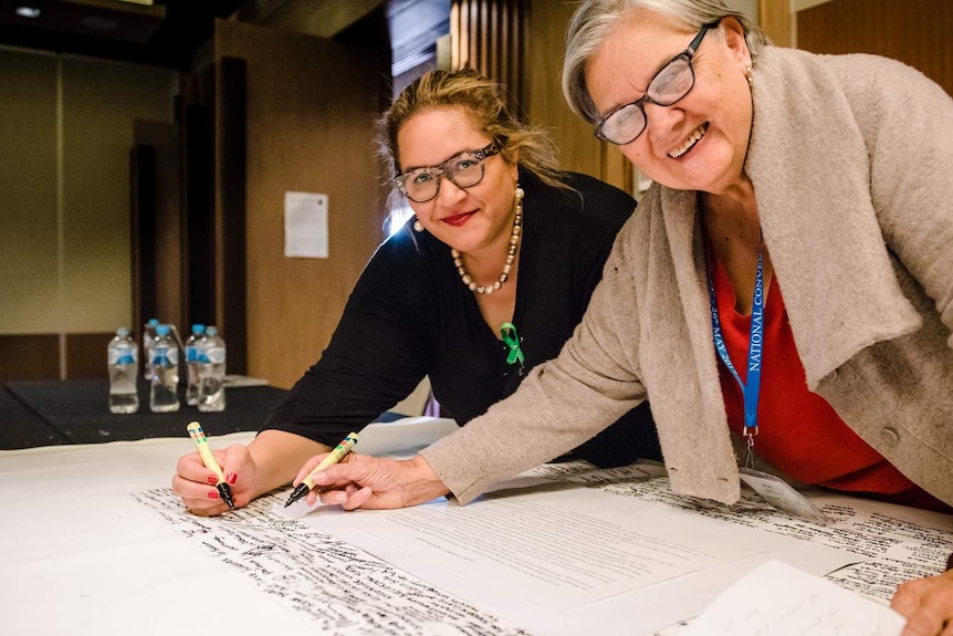 Megan Davis (Left) and Pat Anderson AO bend over a table holding the Uluru Statement from the Heart as she signs the statement.