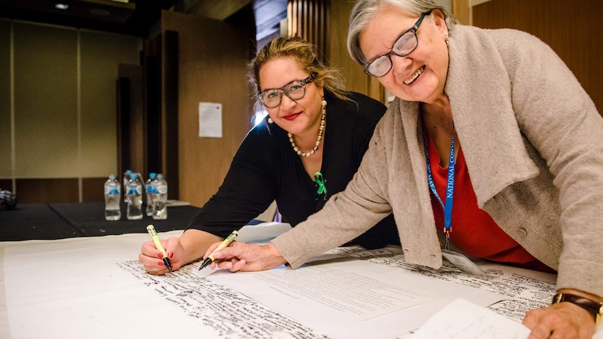 Megan Davis (Left) and Pat Anderson AO bend over a table holding the Uluru Statement from the Heart as she signs the statement.