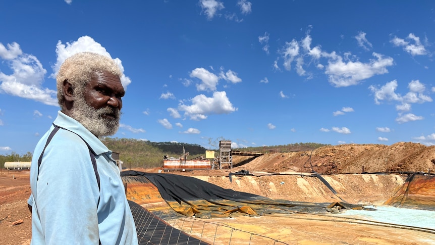 Indigenous man with grey hair stands in front of an old mine site which hasn't been cleaned up