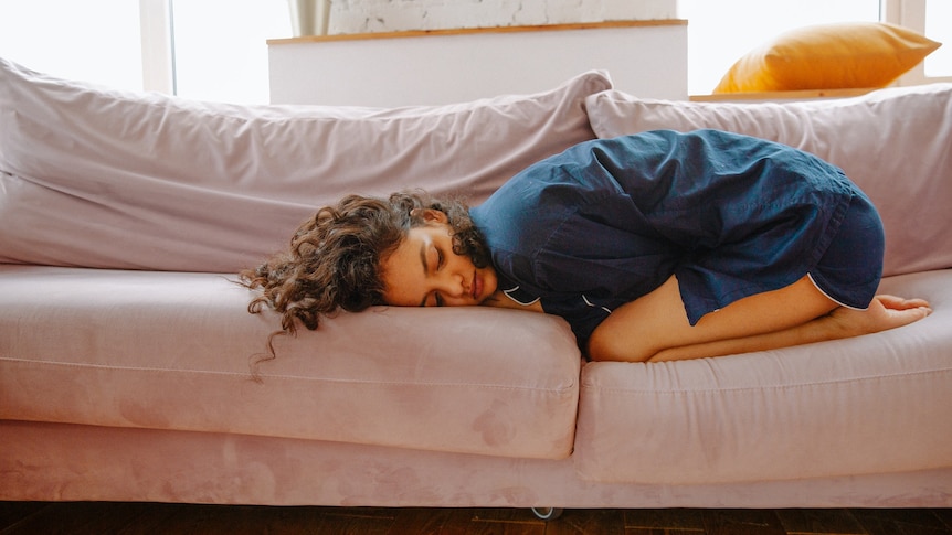 A woman in a blue nightshirt curled up in a ball, in pain, on a pink sofa