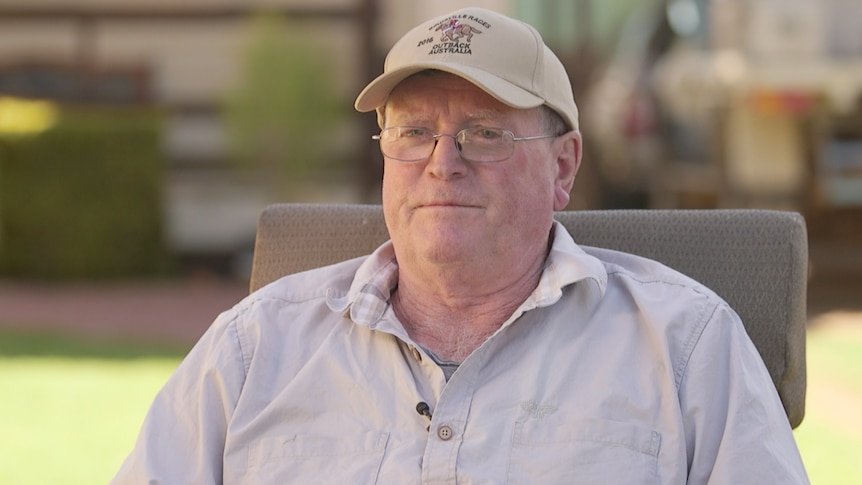 An elderly gentleman in a cap and glasses sits facing the camera, looking off to the side.