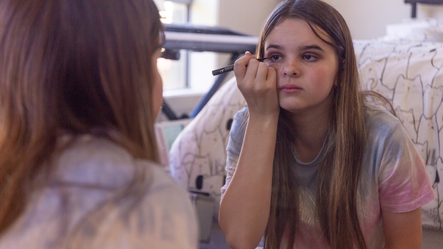 A young white woman sits in front of a bedroom mirror applying eye make up to her face.
