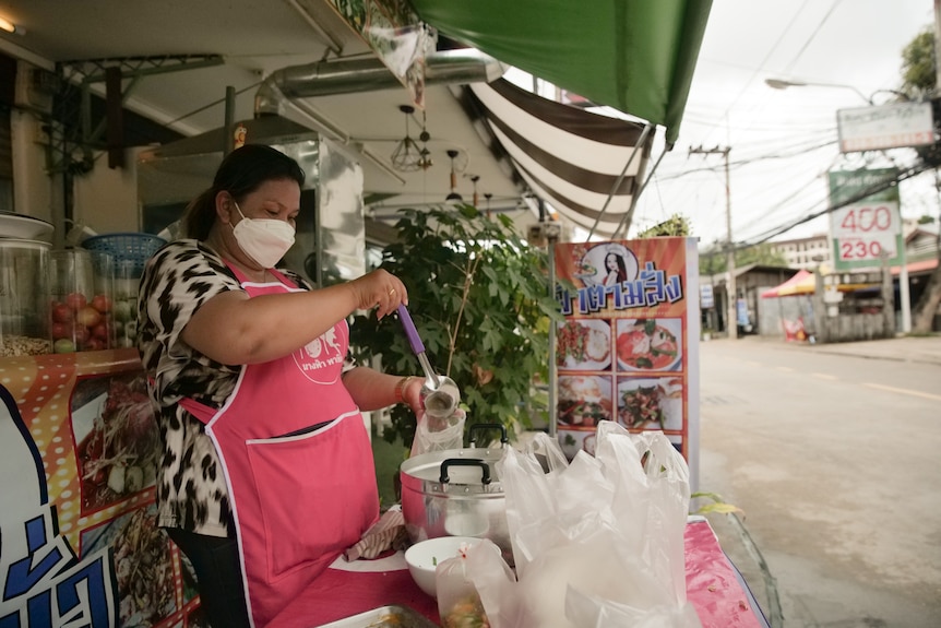 A woman wearing a mask and a pink apron ladles soup into a plastic bag at a shop stall.