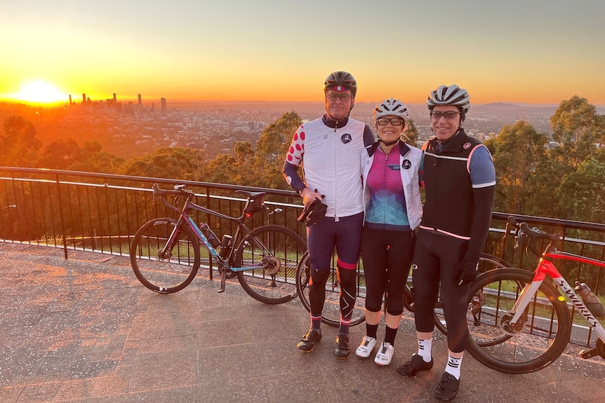 Three bike riders stand with a Brisbane sunrise behind them.