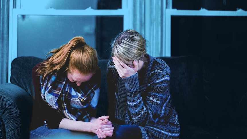 Two women sit on a couch, holding hands, both of their heads bowed in grief supporting each other a sitting with the grief.