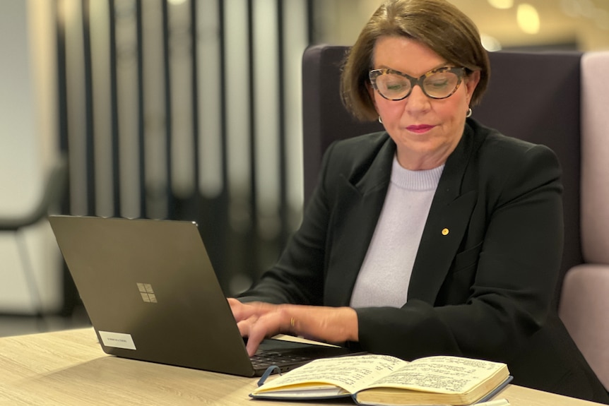 A woman wearing glasses reads a beak while typing on a laptop.